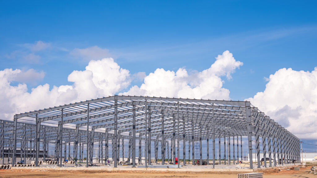 Large metal structure of factory warehouse industry buildings in construction site against clouds on blue sky background